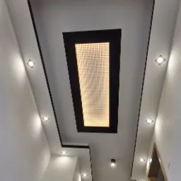 Nowoczesny sufit podwieszany z oświetleniem punktowym i centralnym panelem perforowanym, widoczny z dołu korytarza, z drabiną opartą o ścianę.