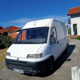 Biały samochód dostawczy Fiat Ducato z widocznymi śladami użytkowania, zaparkowany na betonowym podjeździe przed domem z czerwoną dachówką i błękitnym niebem w tle.