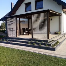 Jasna elewacja domu z drewnianą pergolą i białymi lamelami, taras z roślinami na schodach, zadbany trawnik.