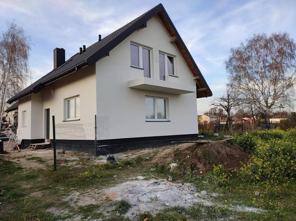 Nowo otynkowany dom jednorodzinny z czarnym dachem i białymi oknami, widoczny z boku, z fragmentem siatki ogrodzeniowej i stertą ziemi na pierwszym planie.