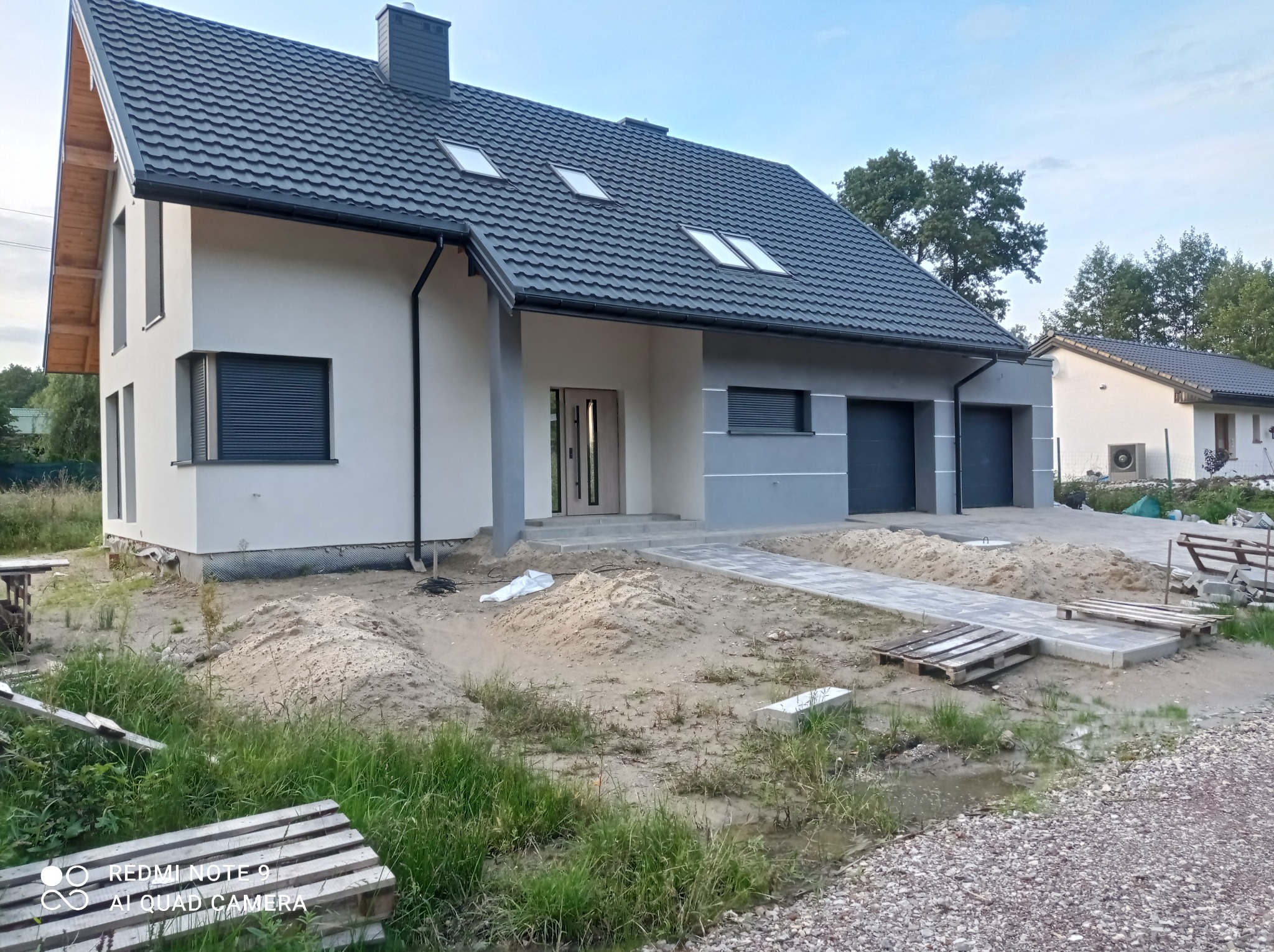 Nowoczesny dom jednorodzinny z szarą elewacją i ciemnoszarą dachówką, widoczne okna dachowe i fragment terenu w trakcie prac wykończeniowych.