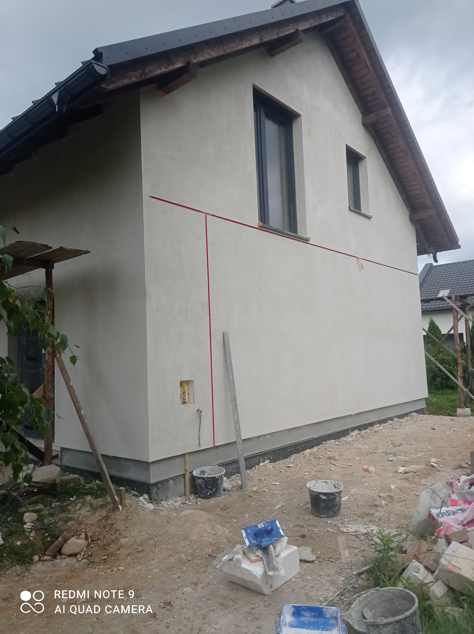 Świeżo otynkowana elewacja domu jednorodzinnego z widocznymi śladami prac budowlanych, w tym wiaderka z zaprawą, rusztowanie i wytyczone linie na ścianie.