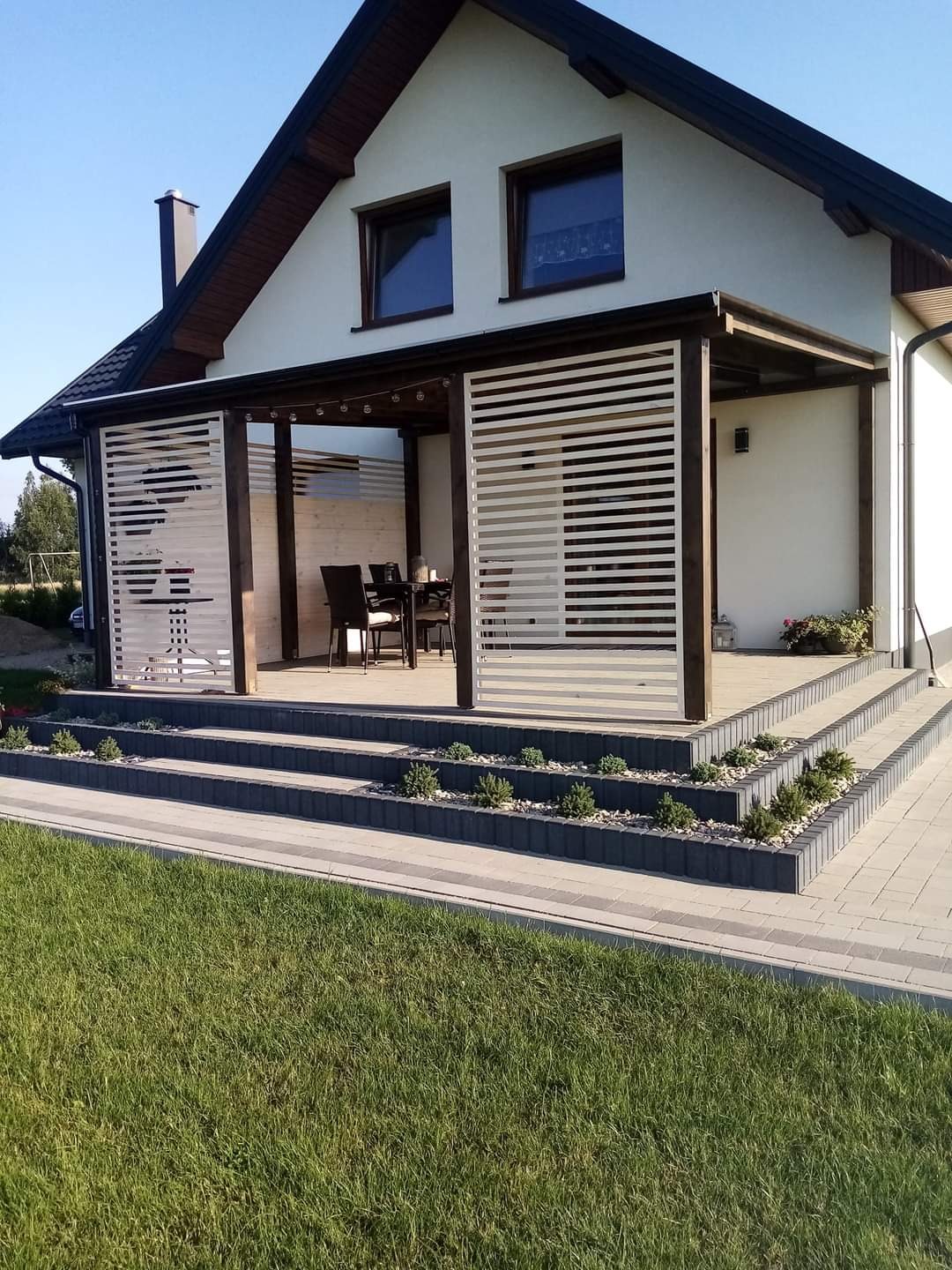 Jasna elewacja domu z drewnianą pergolą i białymi lamelami, taras z roślinami na schodach, zadbany trawnik.