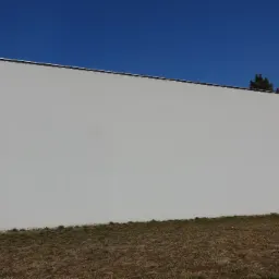 Duża, gładka, pomalowana na biało ściana budynku z widoczną górną krawędzią i fragmentem dachu, na tle błękitnego nieba i suchej trawy u podstawy ściany.