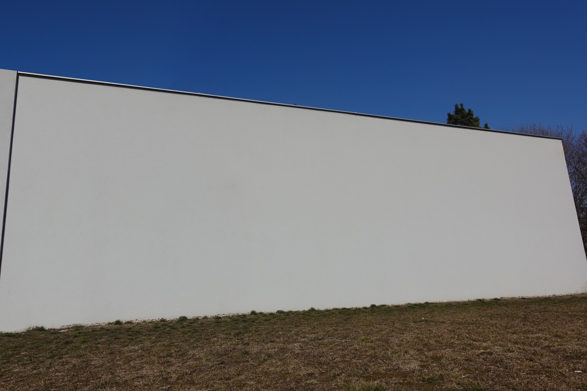 Duża, gładka, pomalowana na biało ściana budynku z widoczną górną krawędzią i fragmentem dachu, na tle błękitnego nieba i suchej trawy u podstawy ściany.