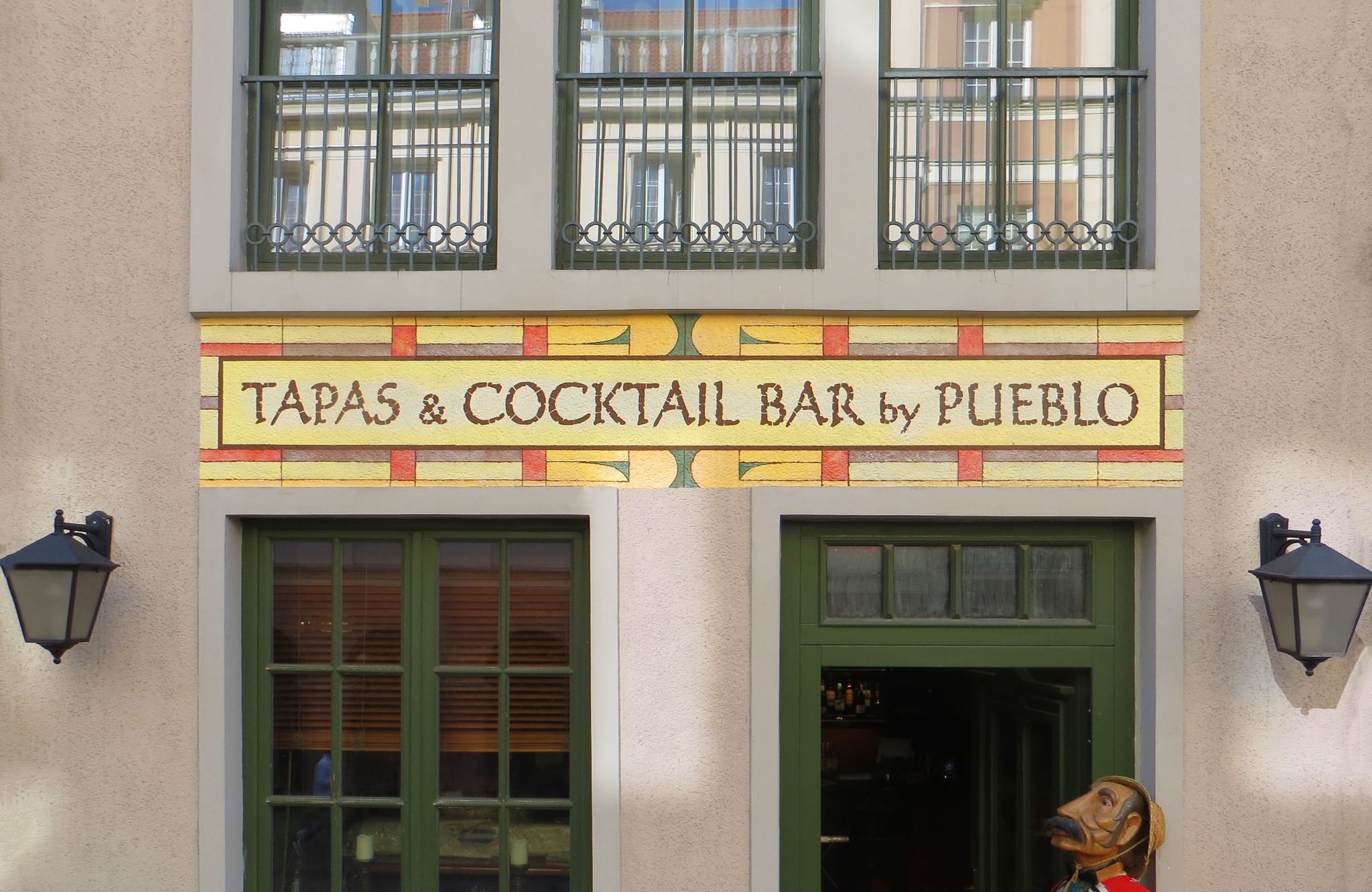 Fasada budynku z napisem 'Tapas & Cocktail Bar by Pueblo', okna z ozdobnymi kratami, zielone drzwi wejściowe i lampy ścienne.