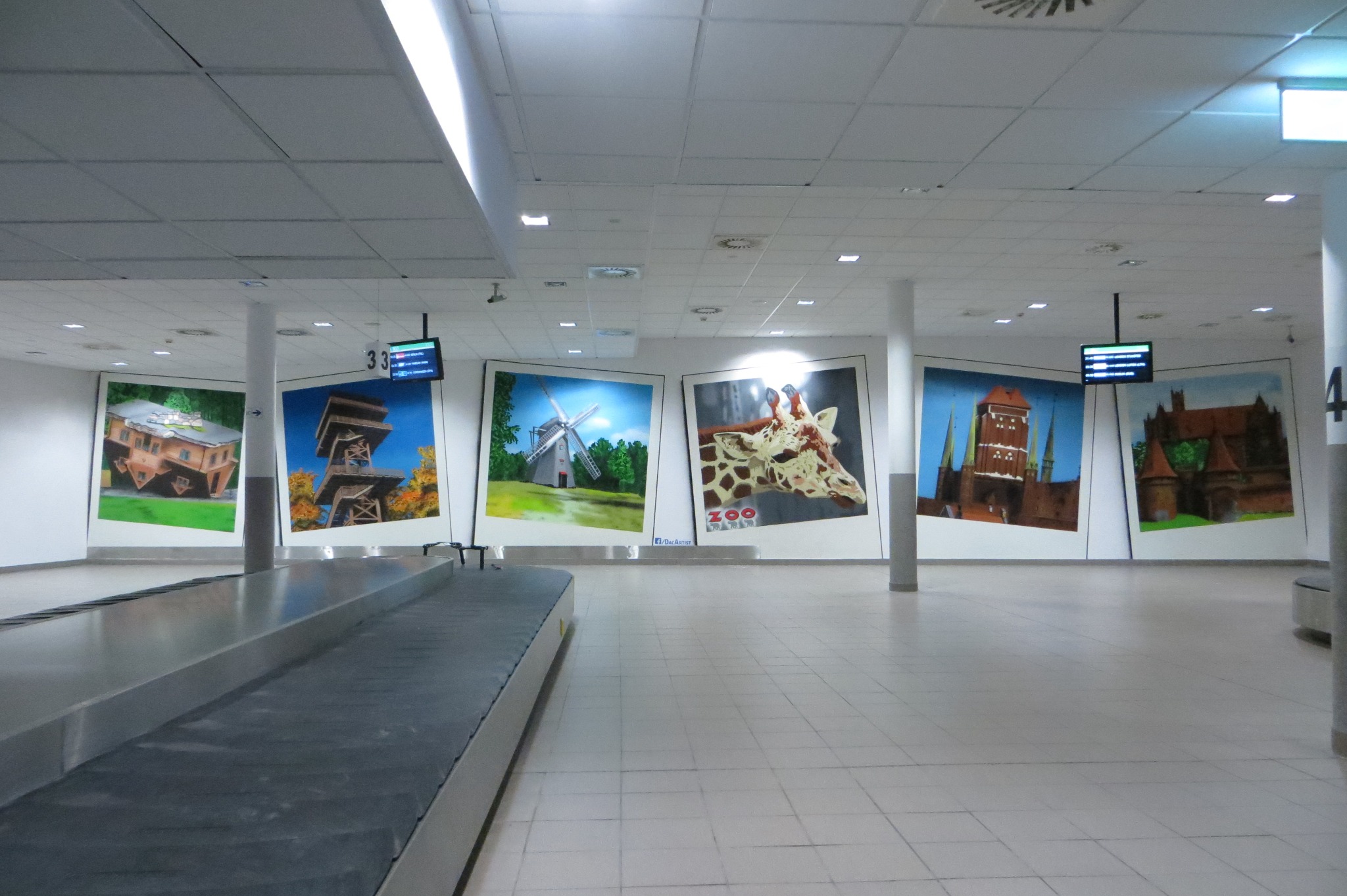Wnętrze hali przylotów z bagażowym pasem transportowym i serią dużych, oprawionych w ramy obrazów na ścianie przedstawiających budynki, wiatrak i żyrafę z zoo.