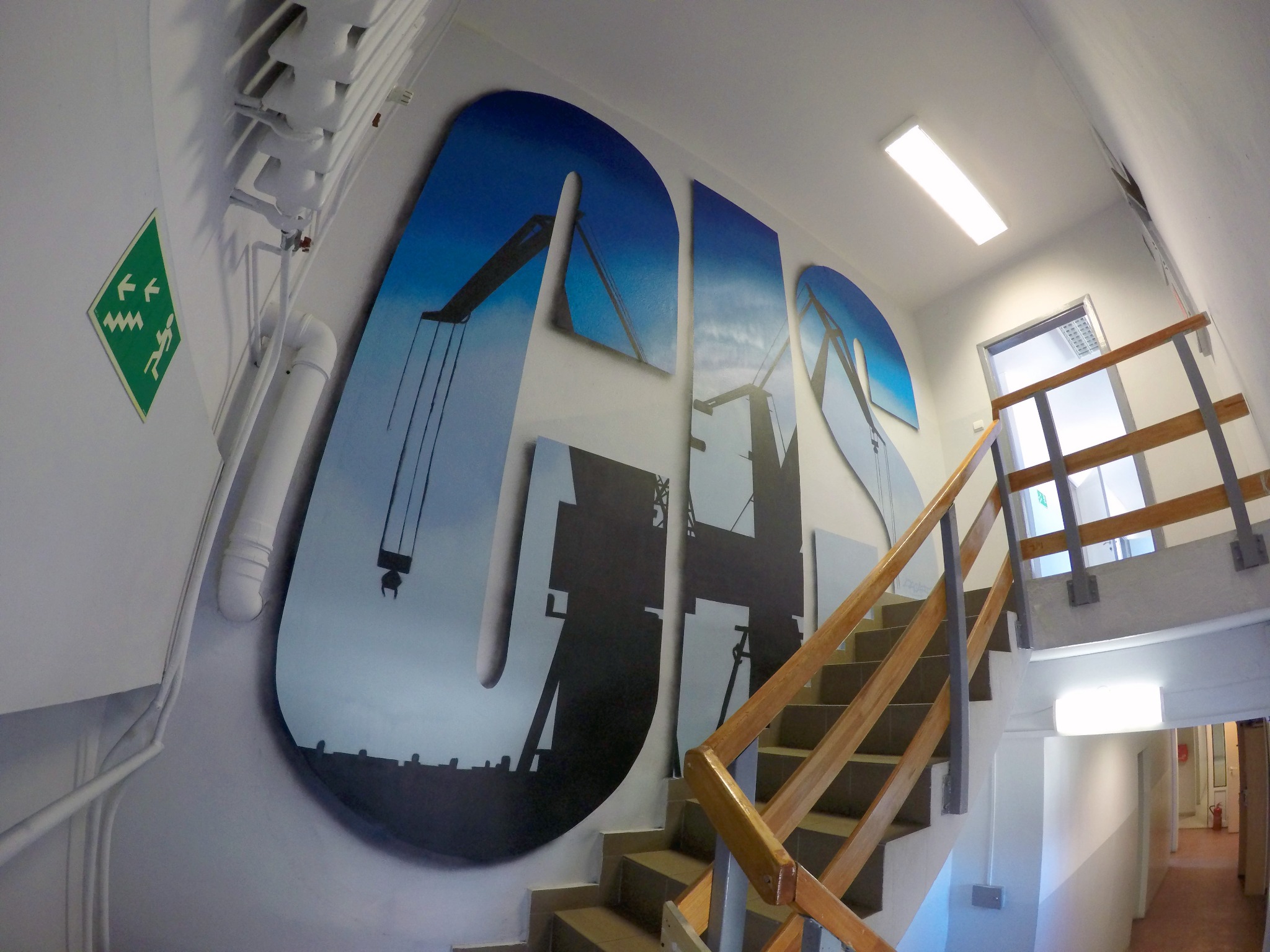 Klatka schodowa z artystycznym malunkiem na ścianie przedstawiającym dźwigi portowe wkomponowane w litery, widoczne rury i oznaczenia ewakuacyjne.