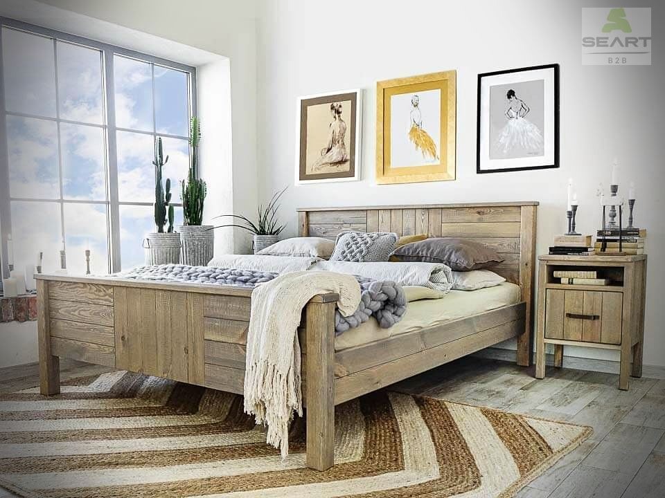 Drewniane łóżko z wezgłowiem z desek, z szafką nocną i dekoracjami, w pokoju z dużym oknem i kaktusami w donicach, na podłodze dywan w pasy.