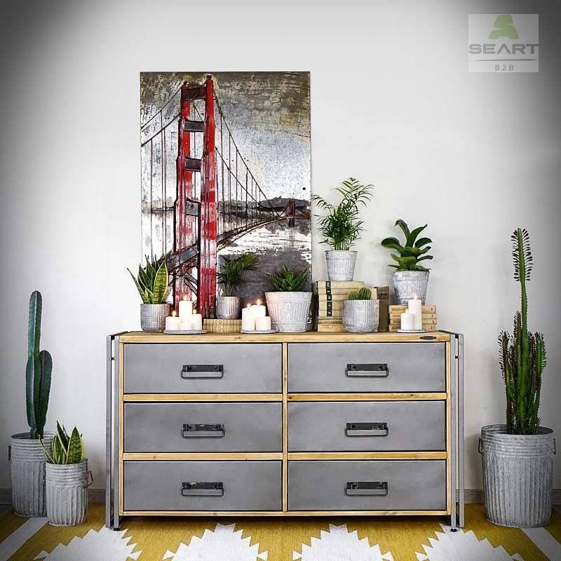 Szara komoda z sześcioma szufladami w stylu industrialnym, na niej rośliny doniczkowe w szarych osłonkach, świece i obraz z motywem mostu Golden Gate, całość na żółto-białym dywanie.