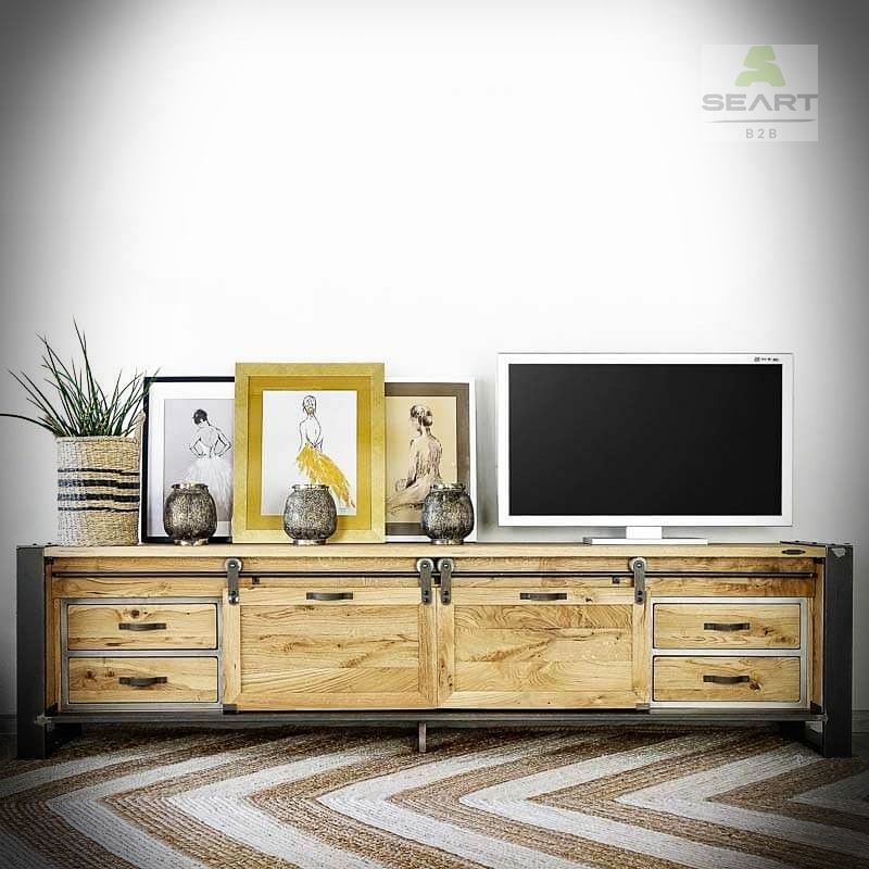 Rustykalna szafka RTV z przesuwanymi drzwiami z litego drewna dębowego, metalowe nogi, dekoracje w postaci ramek z rysunkami i wazonów, na tle biała ściana i dywan w zygzaki.