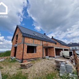 Gwdt Sp. z o.o. - Surowy stan bliźniaka z cegły ceramicznej z czarnym dachem na tle pochmurnego nieba. Widoczne palety i gruz budowlany. Etap budowy przed ociepleniem.