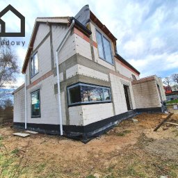 Gwdt Sp. z o.o. - Surowy, dwukondygnacyjny dom murowany w trakcie budowy, z widocznymi oknami z szarymi ramami. Na pierwszym planie ziemia i piasek, w tle drzewa i błękitne niebo.