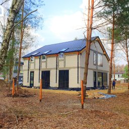 Gwdt Sp. z o.o. - Stan surowy otwarty domu z betonu komórkowego z czarną blachodachówką, widoczny zza drzew na działce. Na dachu okna dachowe.