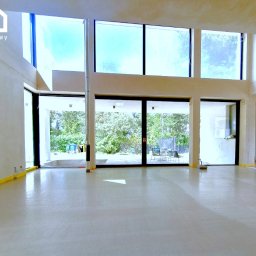 Gwdt Sp. z o.o. - Wnętrze w trakcie wykańczania: duże okna, świeżo wylana posadzka, widoczne przewody elektryczne, taśmy zabezpieczające. Widok na zieleń za oknem.