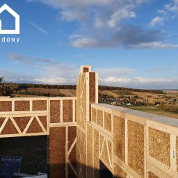 Gwdt Sp. z o.o. - Wznoszenie ścian domu szkieletowego z wypełnieniem ze słomy. Widok na krajobraz z polami i lasami. Konstrukcja z jasnego drewna, wypełnienie słomą. Częściowy widok na niebo.