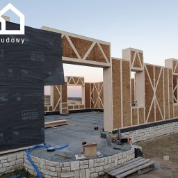 Gwdt Sp. z o.o. - Wznoszenie domu szkieletowego z drewnianą konstrukcją i wypełnieniem z płyt. Widoczne narzędzia i materiały budowlane. Fundament wykończony kamieniem.