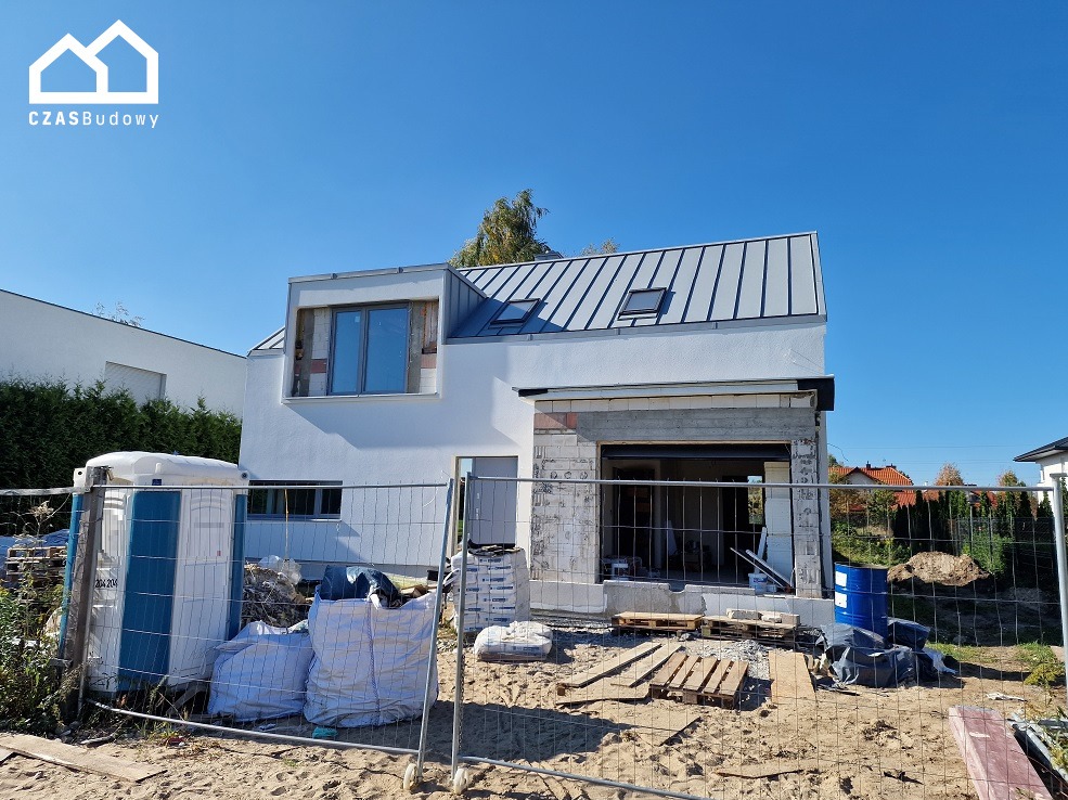 Budowa domu jednorodzinnego w Warszawie: stan surowy otwarty, widoczne materiały budowlane i tymczasowa toaleta. Dach metalowy, okna wstawione.