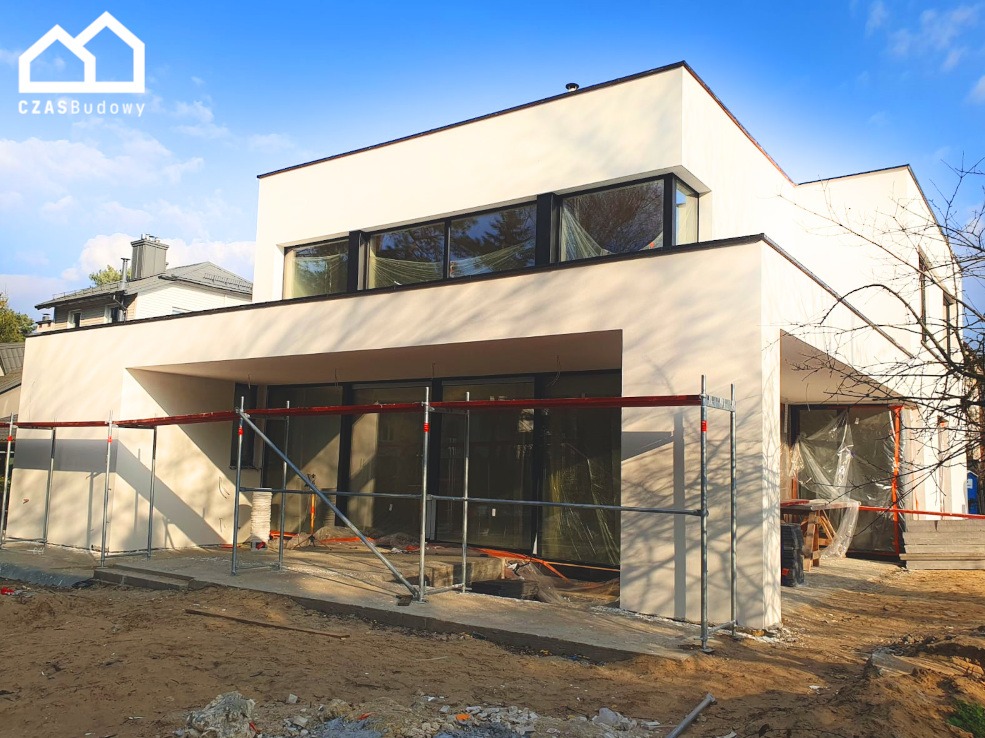 Nowoczesny dom w trakcie wykończenia elewacji, widoczne rusztowania i folia ochronna na oknach, jasna elewacja, błękitne niebo.