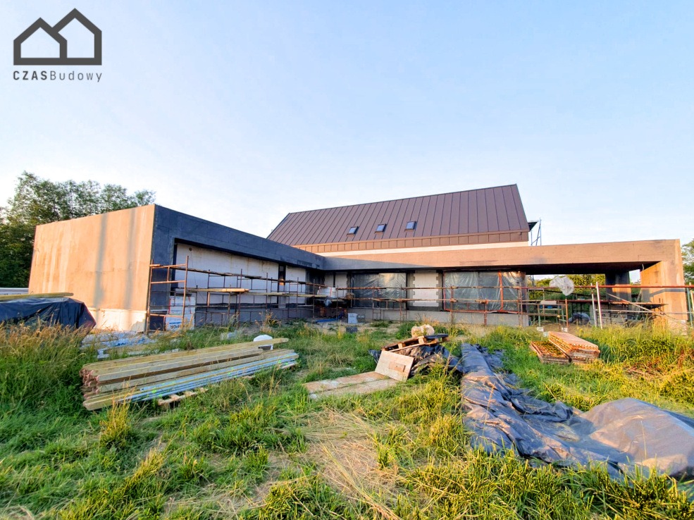 Surowy stan domu jednorodzinnego z ciemnym dachem i oknami, w trakcie budowy, z widocznymi rusztowaniami i materiałami budowlanymi na zarośniętej działce.