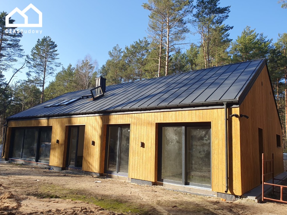 Nowoczesny dom szkieletowy z drewnianą elewacją i grafitowym dachem na tle sosnowego lasu. Widoczne duże przesuwne okna i czarny komin.