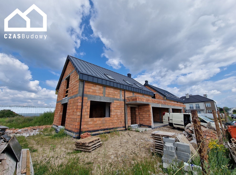 Surowy stan bliźniaka z cegły ceramicznej z czarnym dachem na tle pochmurnego nieba. Widoczne palety i gruz budowlany. Etap budowy przed ociepleniem.