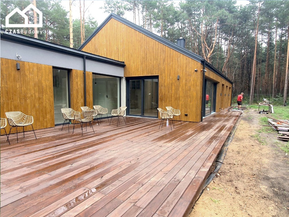 Nowoczesny dom z drewnianą elewacją i tarasem, otoczony lasem. Widoczne krzesła na tarasie i częściowo zrealizowana zabudowa tarasu z boku budynku.