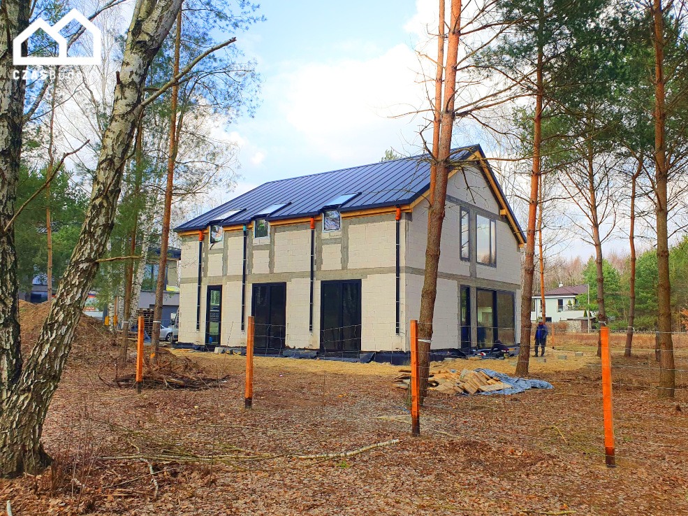 Stan surowy otwarty domu z betonu komórkowego z czarną blachodachówką, widoczny zza drzew na działce. Na dachu okna dachowe.