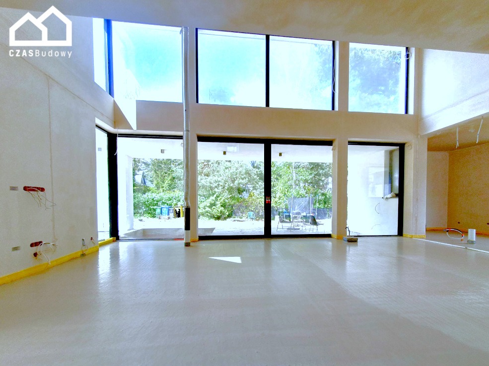 Wnętrze w trakcie wykańczania: duże okna, świeżo wylana posadzka, widoczne przewody elektryczne, taśmy zabezpieczające. Widok na zieleń za oknem.