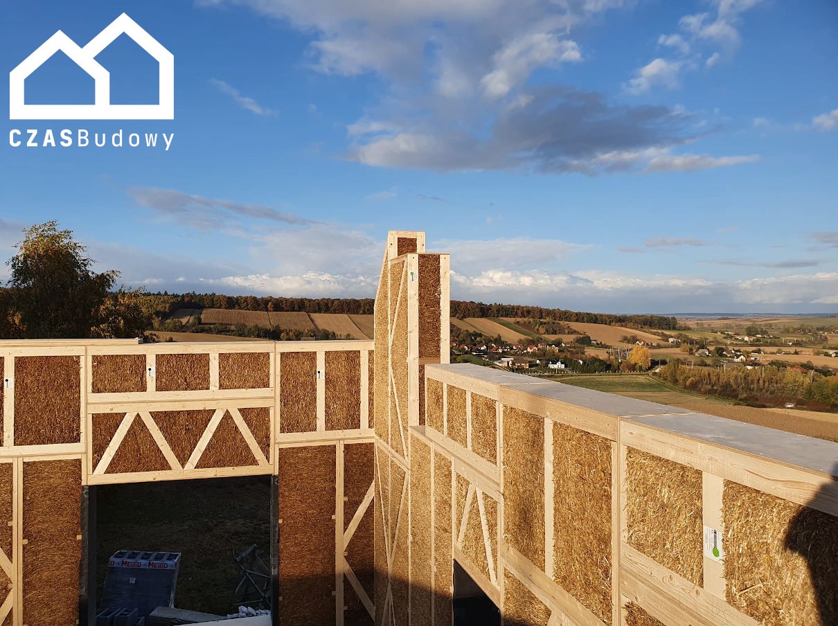 Wznoszenie ścian domu szkieletowego z wypełnieniem ze słomy. Widok na krajobraz z polami i lasami. Konstrukcja z jasnego drewna, wypełnienie słomą. Częściowy widok na niebo.