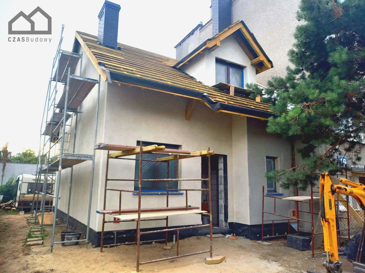 Elewacja domu jednorodzinnego w trakcie prac wykończeniowych. Widoczne rusztowania, surowe drewno na dachu i świeżo nałożony tynk. Żółty mini-koparka w tle.