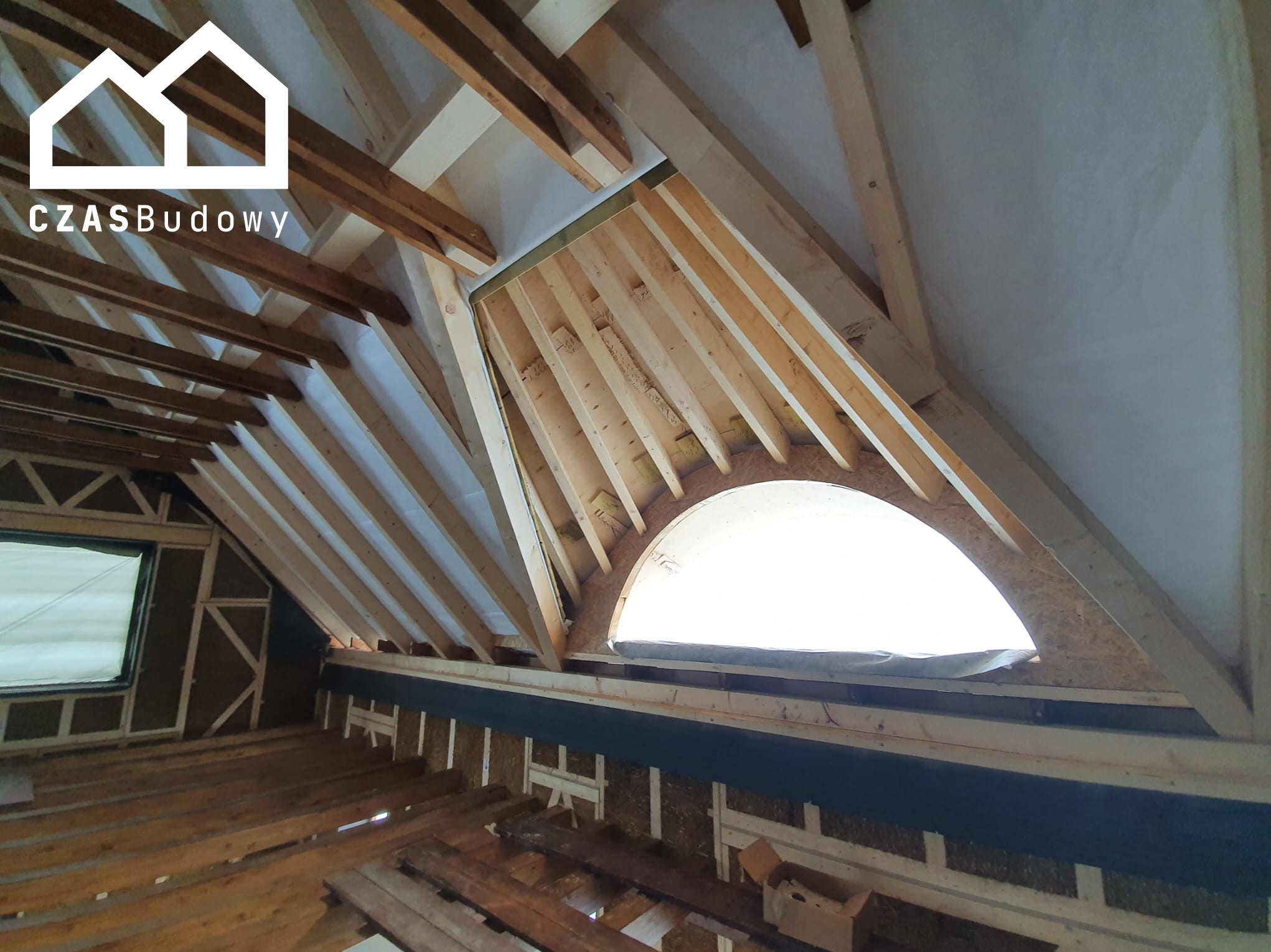 Konstrukcja dachu z drewnianymi belkami i okrągłym oknem, widoczne ocieplenie ścian. Etap prac wykończeniowych poddasza z logo firmy w rogu.