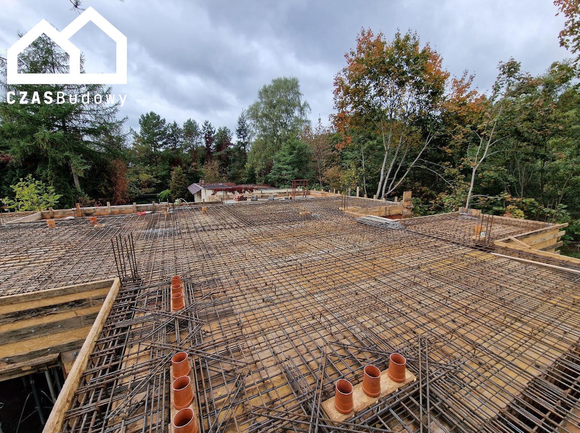 Zbrojenie stropu domu jednorodzinnego z widocznymi rurami kanalizacyjnymi, przygotowane do zalania betonem. Widok z góry na tle zieleni.