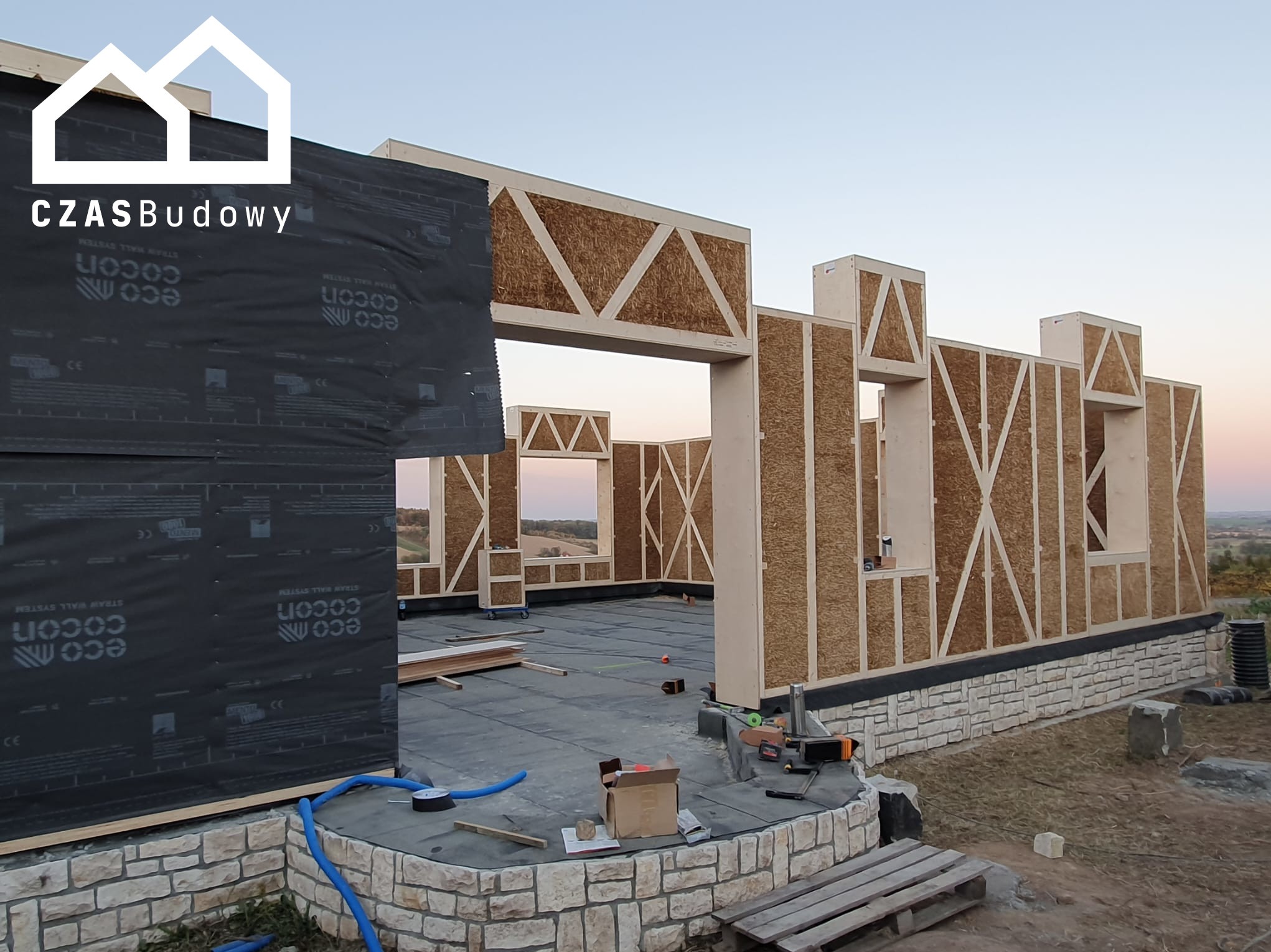 Wznoszenie domu szkieletowego z drewnianą konstrukcją i wypełnieniem z płyt. Widoczne narzędzia i materiały budowlane. Fundament wykończony kamieniem.