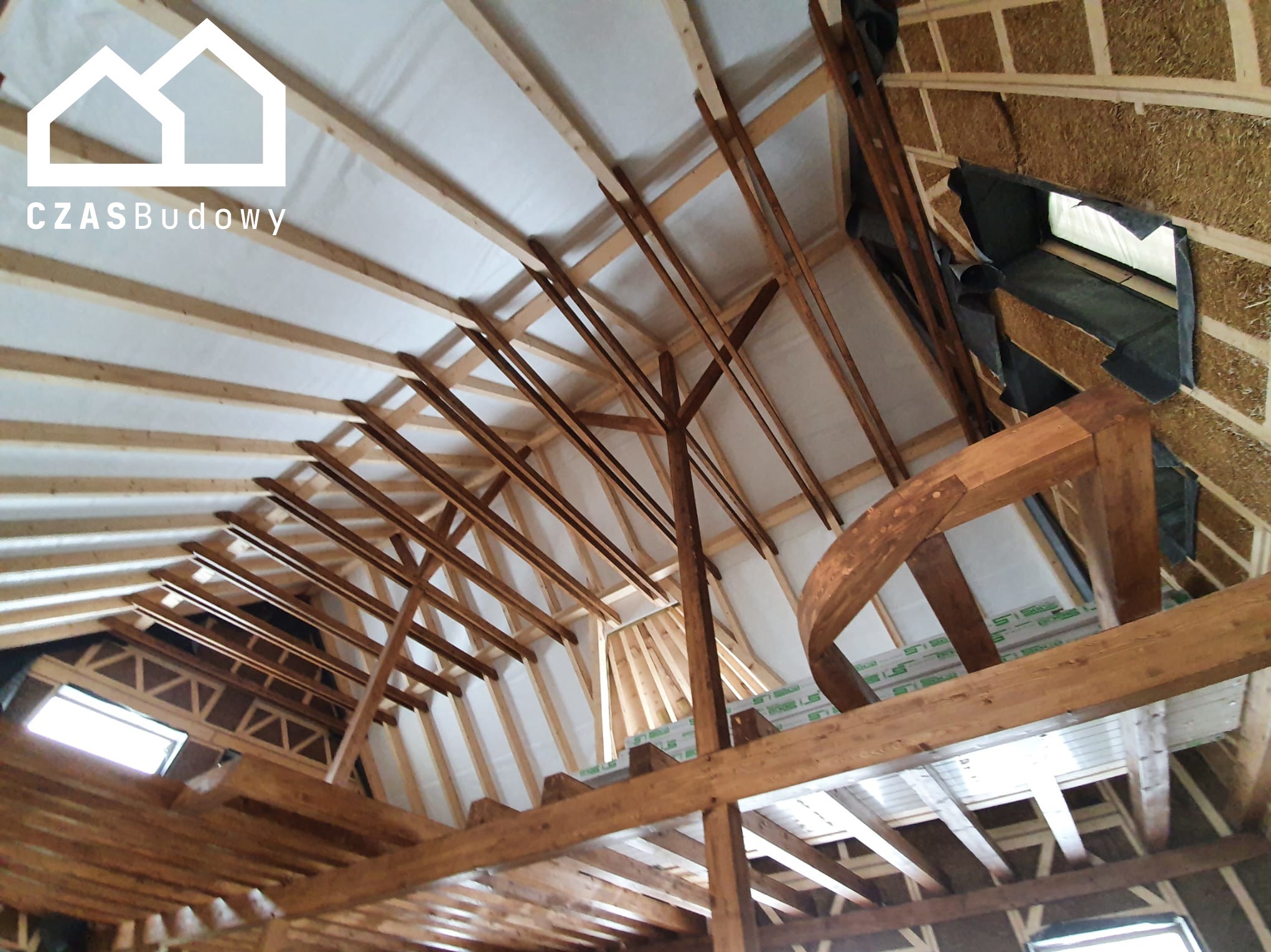 Wnętrze domu w trakcie budowy: widok na drewnianą konstrukcję dachu, izolację i fragment ściany z wypełnieniem ze słomy. Ujęcie od dołu, z widocznymi oknami.