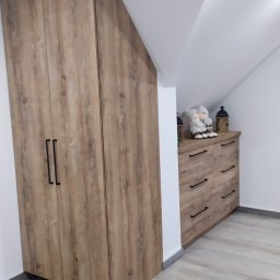 Molenda Meble Na Wymiar - Drewniana szafa z czarnymi uchwytami, dopasowana do skosu dachu, obok komoda z dekoracjami w pokoju na poddaszu.