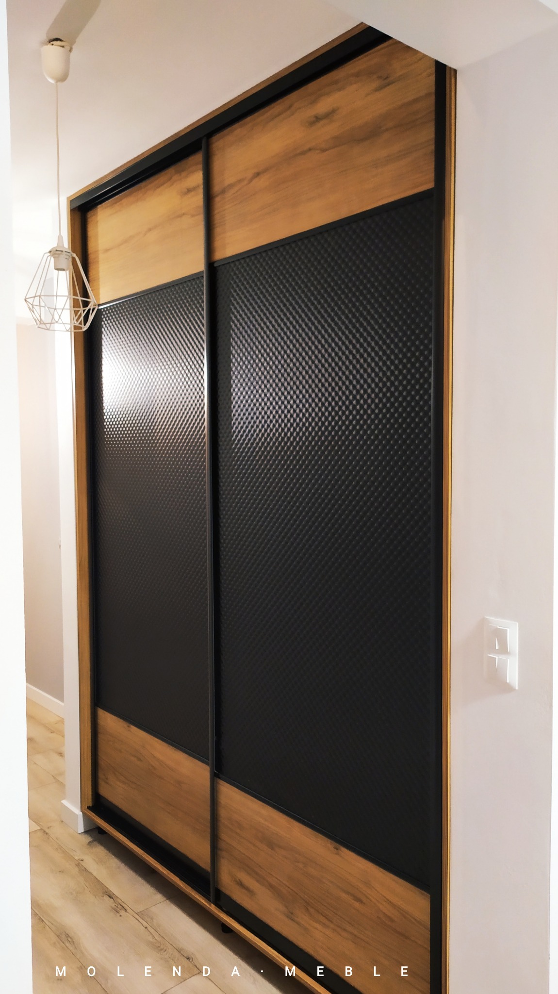 Nowoczesna szafa przesuwna z drewnianymi elementami i czarnymi panelami z fakturą, oświetlona wiszącą lampą drucianą.