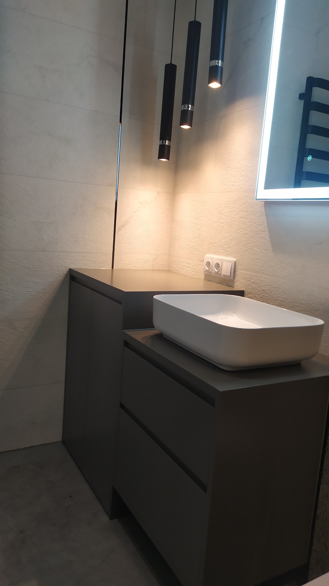 Nowoczesna łazienka z szarą szafką pod umywalkę nablatową, oświetlona wiszącymi lampami tubami i lustrem z podświetleniem LED.