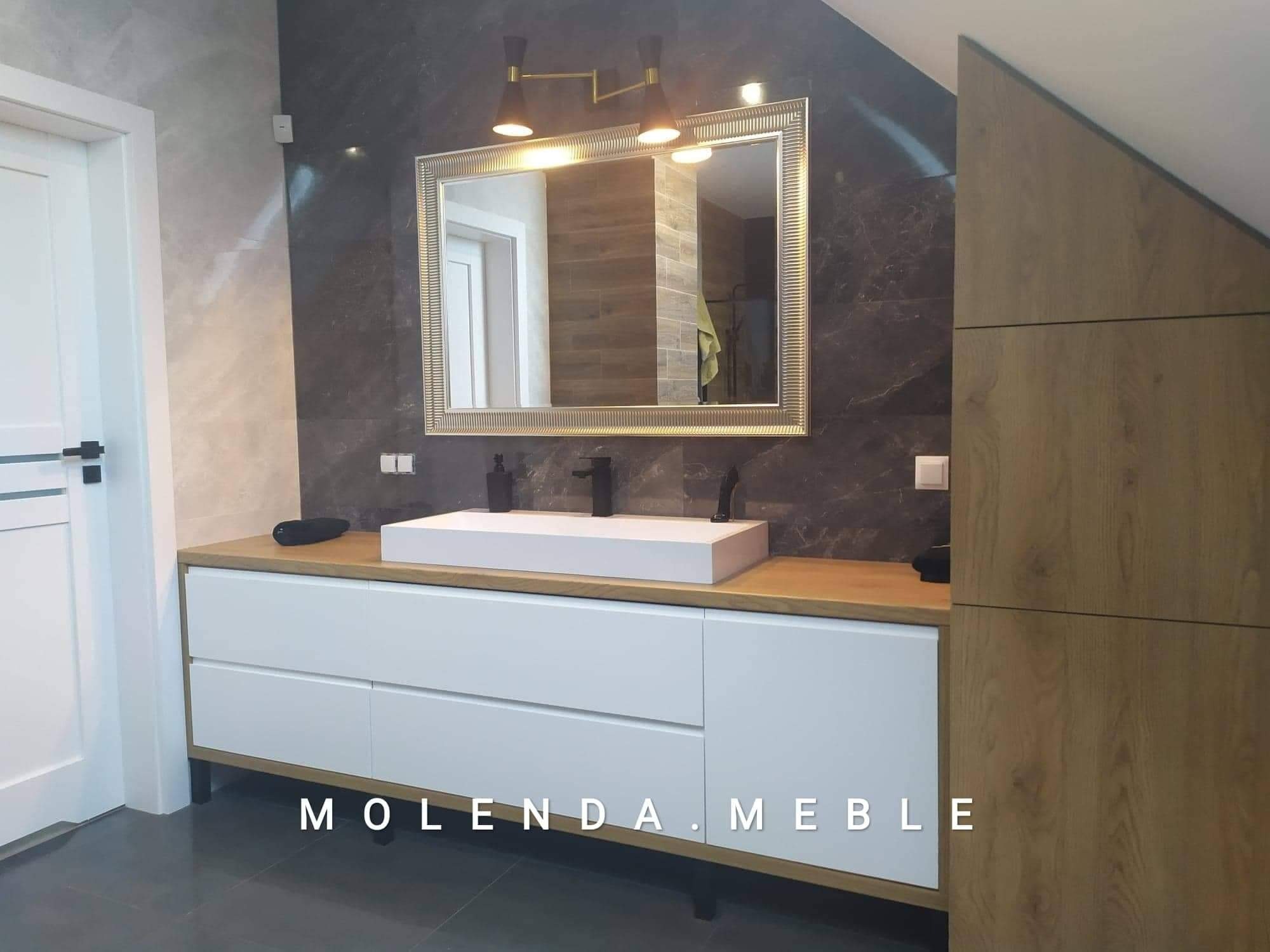 Nowoczesna łazienka z białą szafką pod umywalkę z drewnianym blatem, lustrem w złotej ramie i marmurowymi płytkami na ścianie.