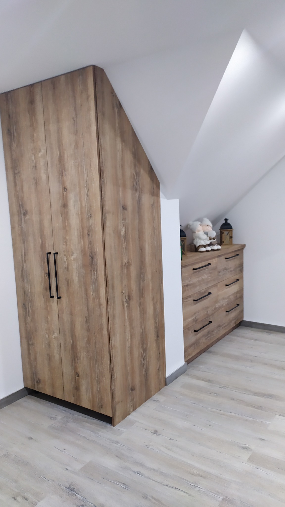 Drewniana szafa z czarnymi uchwytami, dopasowana do skosu dachu, obok komoda z dekoracjami w pokoju na poddaszu.