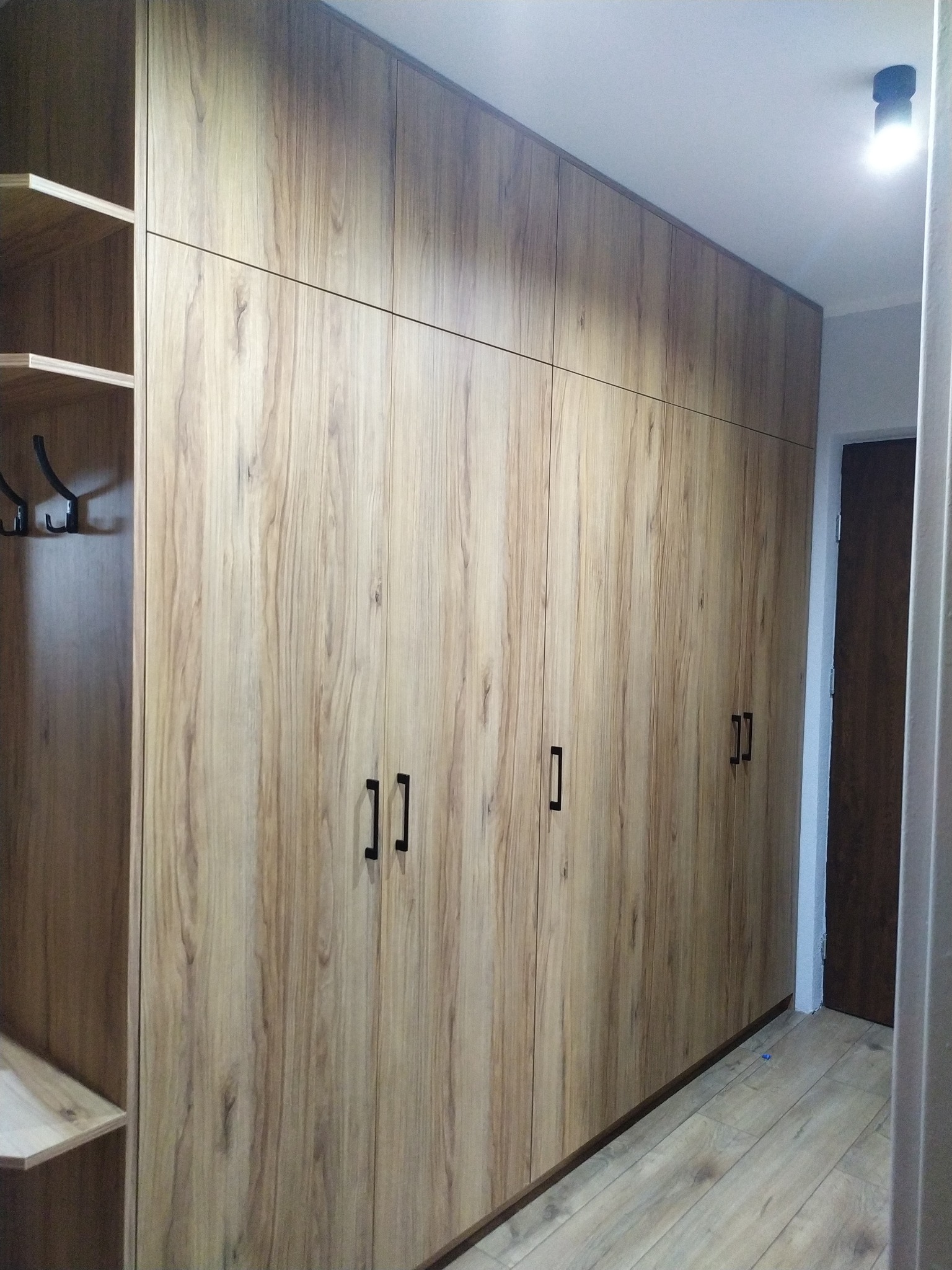 Wysoka zabudowa szafy z frontami imitującymi drewno, z czarnymi, prostokątnymi uchwytami, po lewej stronie otwarta garderoba z wieszakami.