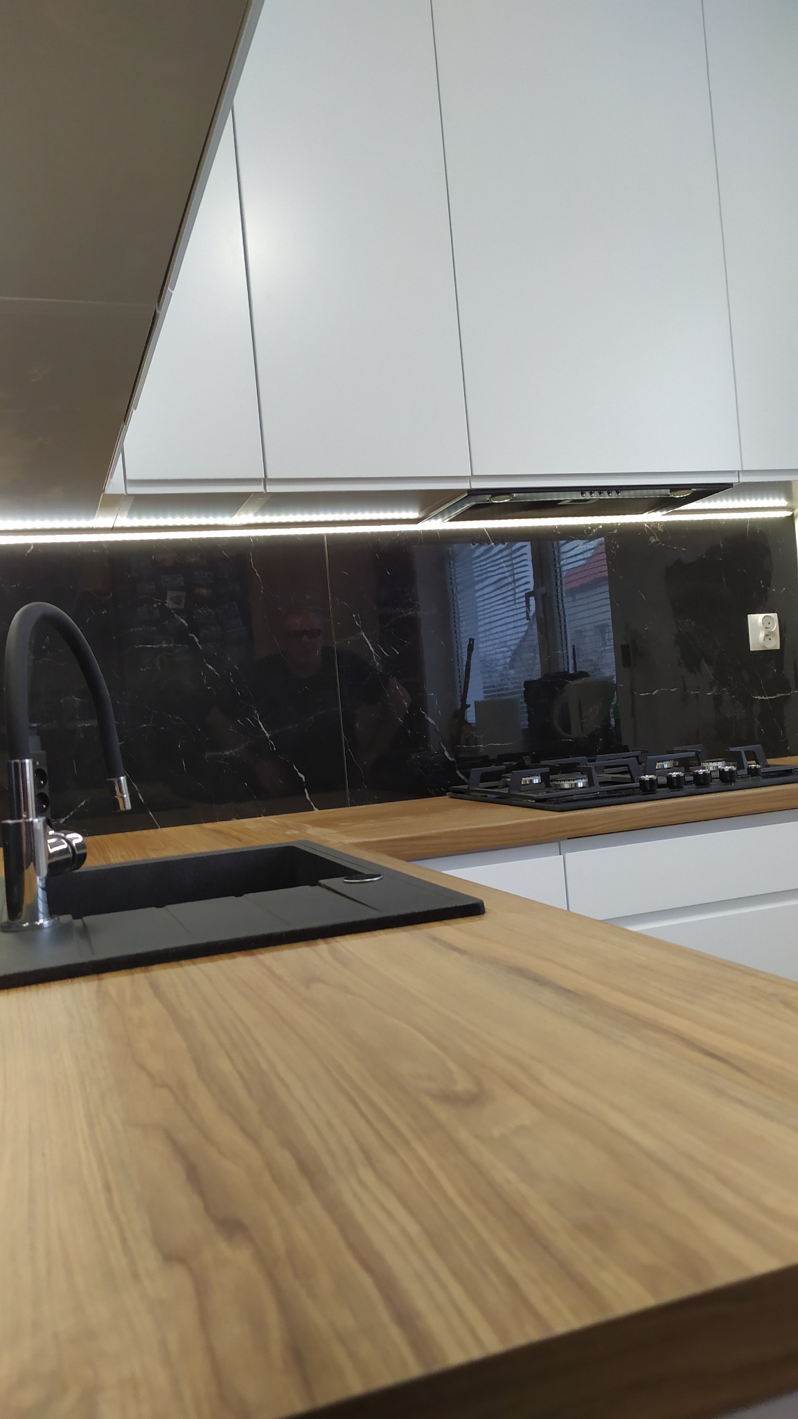 Nowoczesna kuchnia: drewniany blat, czarny zlew i bateria, marmurowe panele ścienne, białe szafki górne z oświetleniem LED, płyta gazowa.