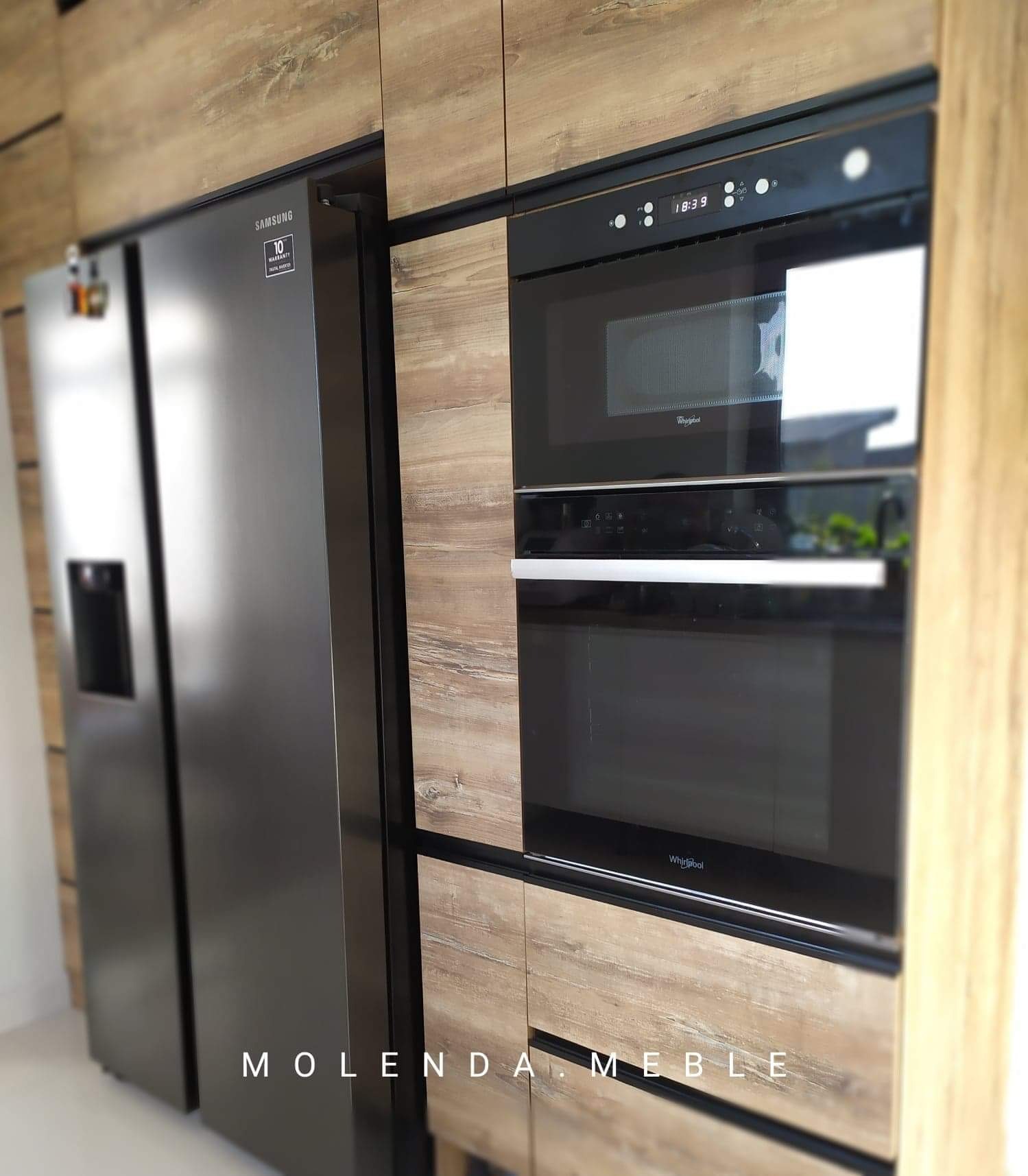 Nowoczesna zabudowa kuchenna z drewnopodobnymi frontami, czarnymi akcentami, wbudowaną lodówką side-by-side Samsung i piekarnikiem z mikrofalą Whirlpool.