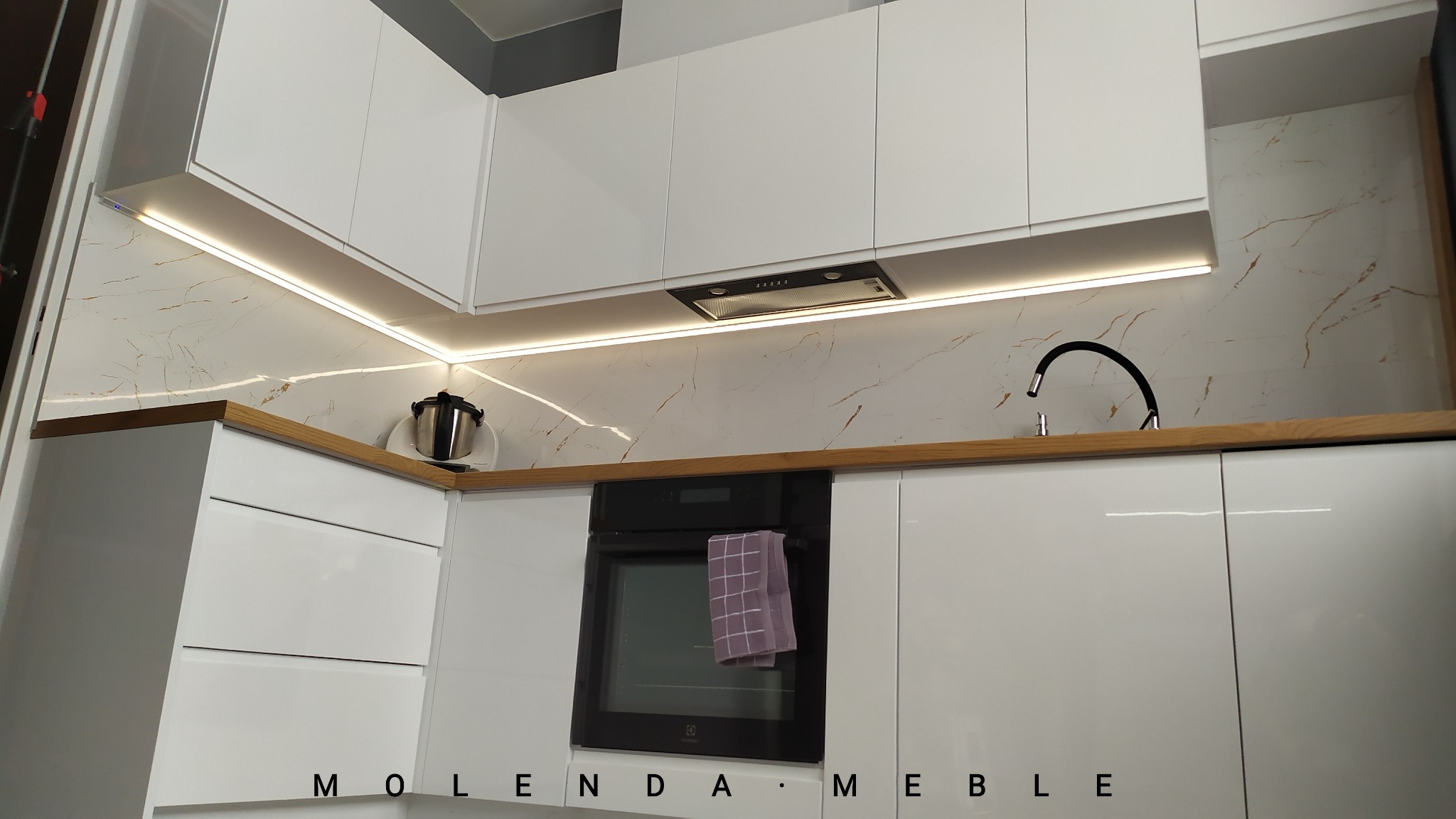 Nowoczesna biała kuchnia z drewnianym blatem, podświetleniem LED i marmurowym wykończeniem ściany, z czarną armaturą i piekarnikiem z wiszącym ręcznikiem.