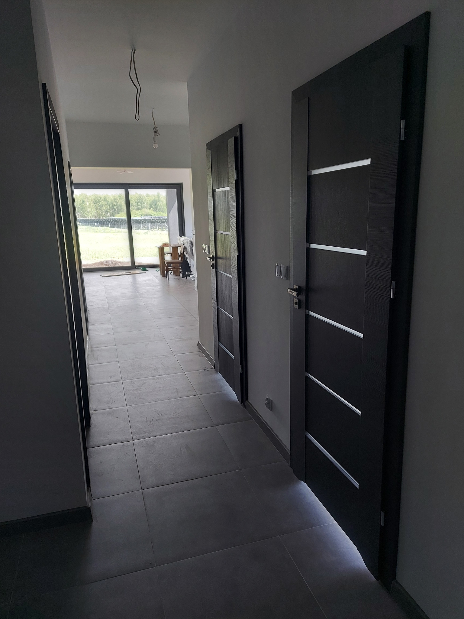 Nowoczesny korytarz z trzema ciemnymi drzwiami z poziomymi, jasnymi wstawkami, szare płytki podłogowe, widok na salon z dużym oknem tarasowym i niezakończone prace wykończeniowe.
