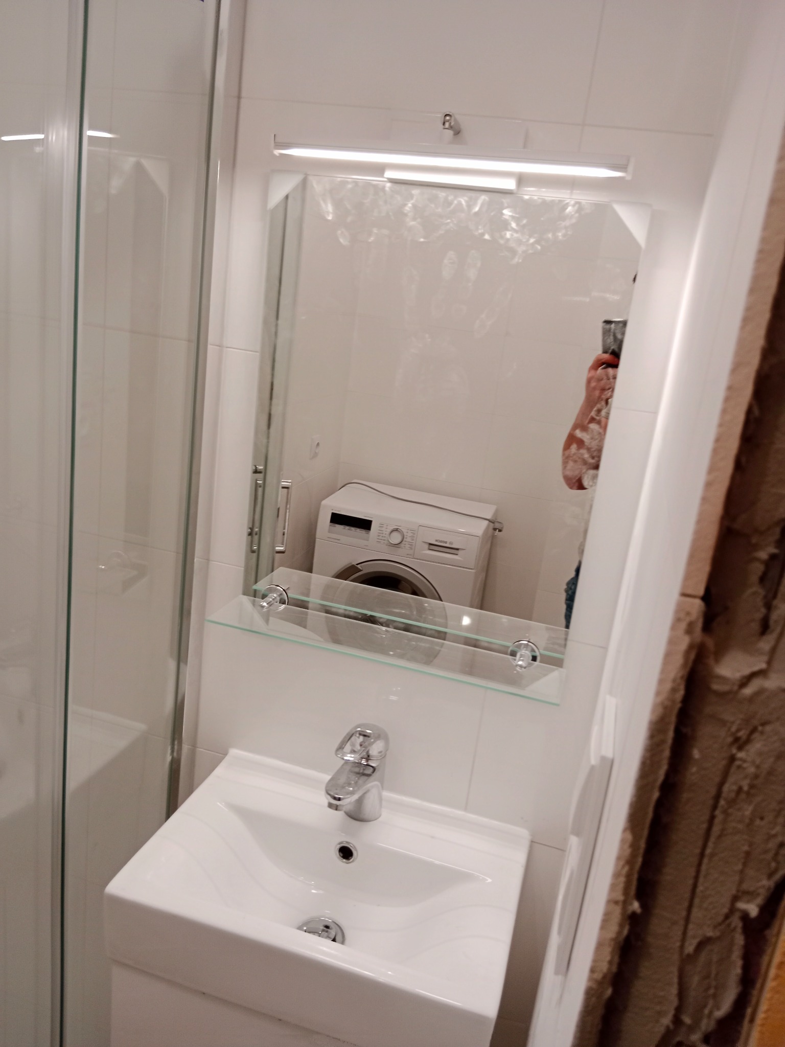 Mała, biała łazienka z kabiną prysznicową, umywalką z szafką i pralką widoczną w lustrze, fragment ściany w trakcie remontu.