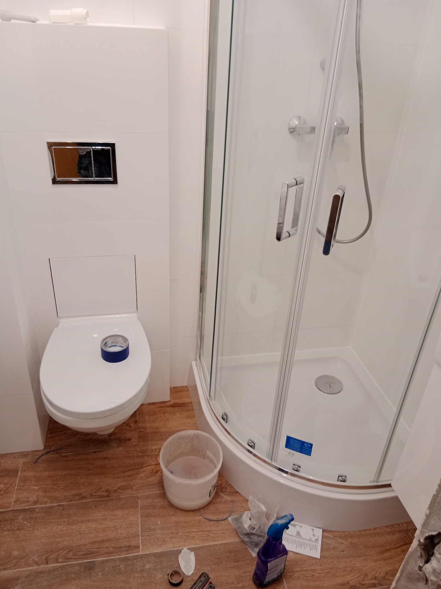 Biała, nowoczesna łazienka w trakcie prac wykończeniowych: kabina prysznicowa z przezroczystymi drzwiami, podwieszana toaleta z niebieską taśmą zabezpieczającą, drewnopodobne płytki na podłodze...