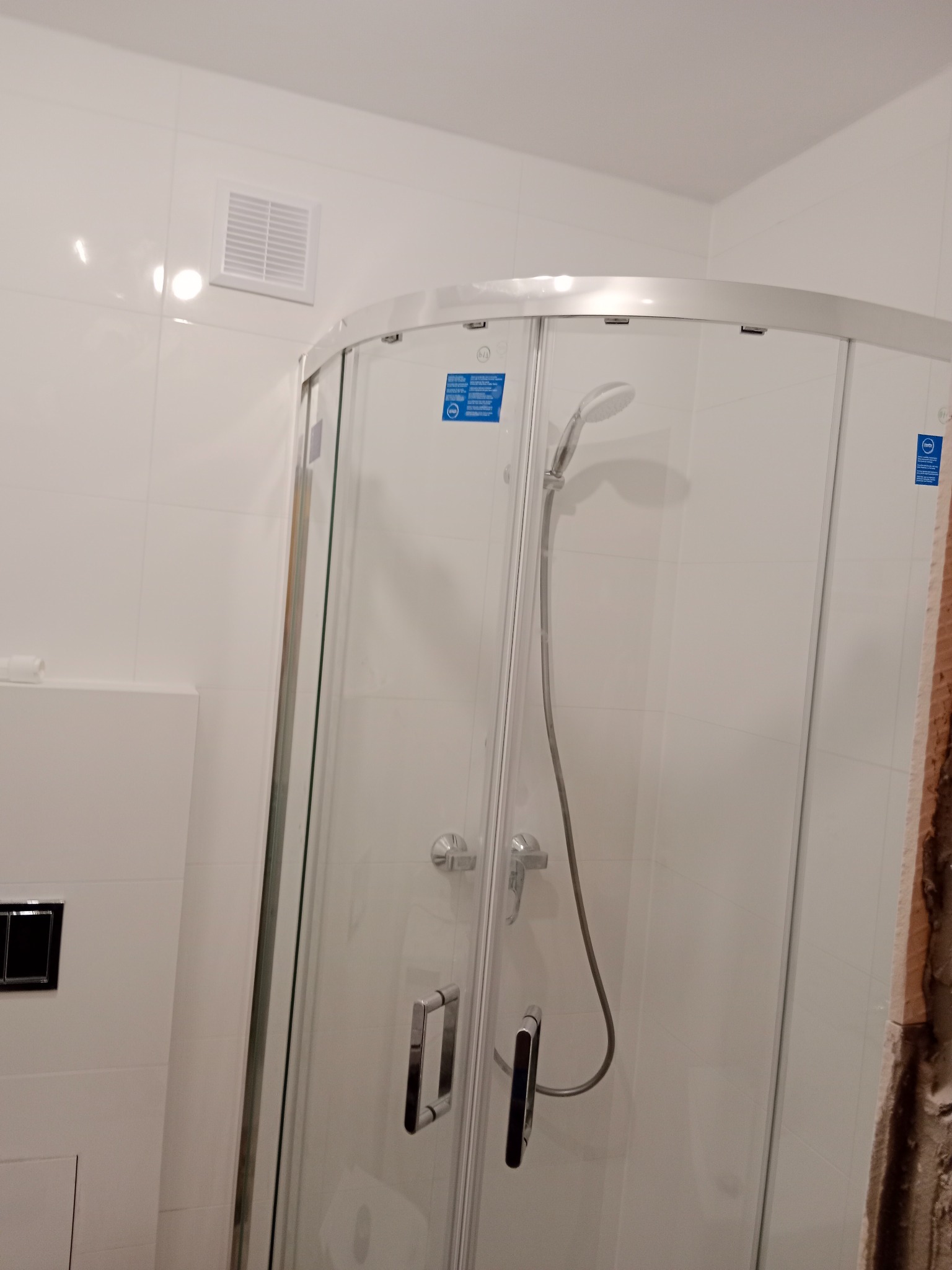 Narożna kabina prysznicowa z chromowanymi uchwytami w łazience z białymi płytkami, widoczny fragment nieukończonej ściany z prawej strony.