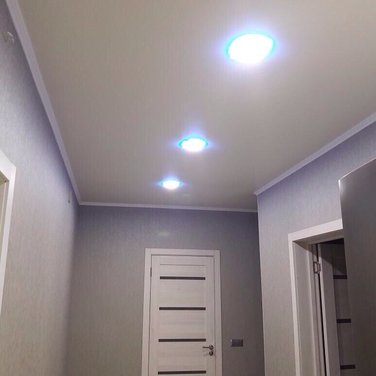 Korytarz z białym sufitem podwieszanym i trzema okrągłymi lampami LED z niebieską poświatą, ściany w odcieniach szarości z delikatnym wzorem, białe drzwi z poziomymi panelami.