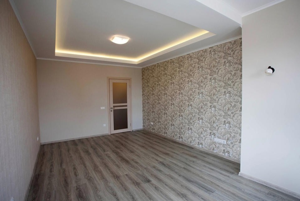 Pusty pokój po remoncie z panelami podłogowymi w odcieniach szarości, ścianą w beżowej tapecie we wzory, podwieszonym sufitem z oświetleniem LED i drzwiami z szybą.